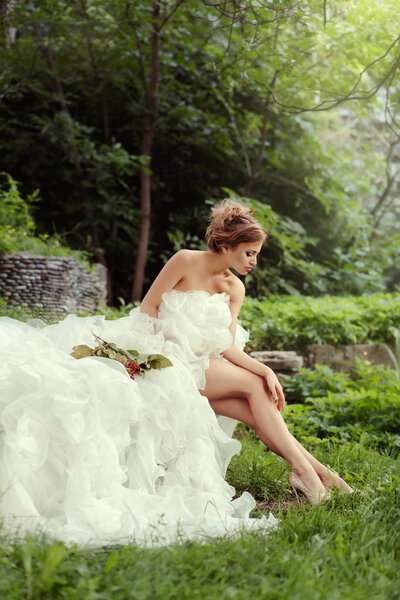 Beautiful woman bride looking at her long legs in nature in the forest sitting on the grass.