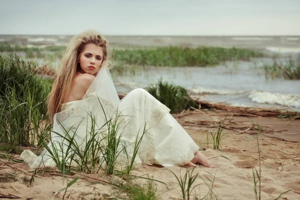 Mooi meisje in een witte jurk zit aan de kust onder de rukwinden van een koude wind. — Stockfoto