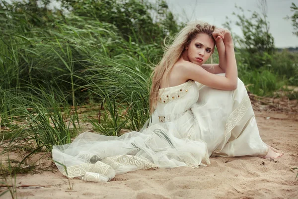 Hermosa chica en un vestido blanco está sentado en la playa y mirando hacia atrás . Imágenes De Stock Sin Royalties Gratis