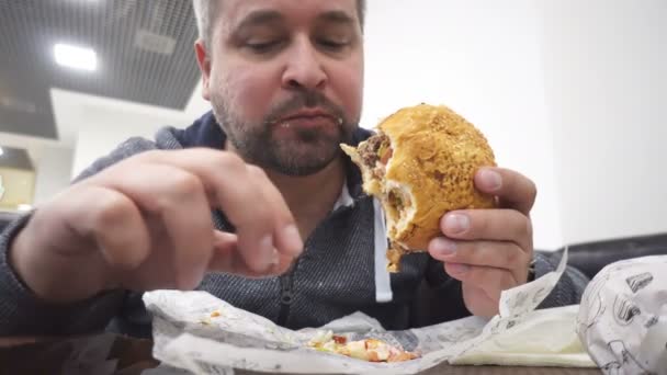 Голодный человек ест большой гамбургер в кафе быстрого питания — стоковое видео