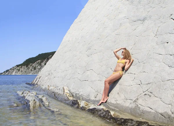 Beautiful blonde posing leaning against rock near ocean coast — Stock Photo, Image