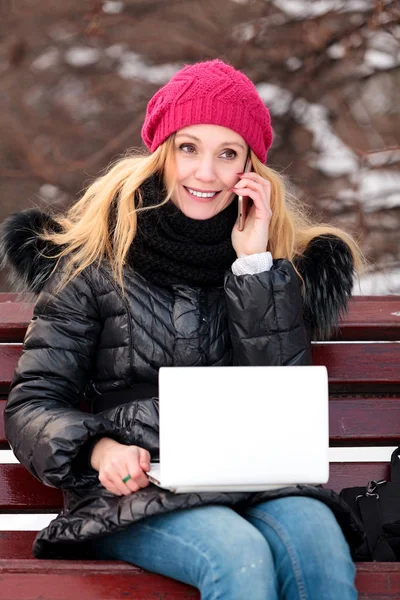 Mosolygó nő ül egy padon a parkban, beszél a mobiltelefonján, és használ laptop portréja. — Stock Fotó
