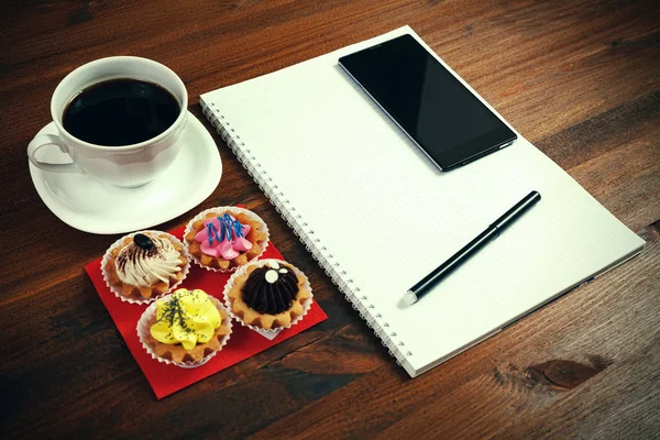 Witte koffiekopje met notebook, vier cupcakes, smartphone en pen op een houten tafel. — Stockfoto