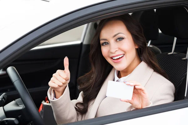 Schönes junges Mädchen sitzt im weißen Auto und zeigt eine leere weiße Karte und Daumen nach oben Handzeichen — Stockfoto