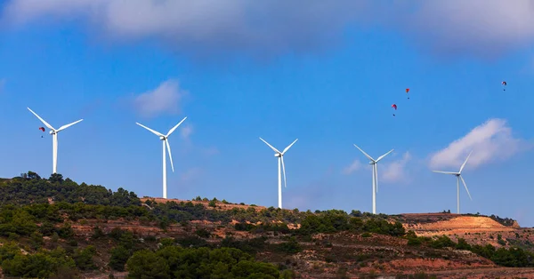 青い空と空気中の4つのパラグライダーに対して丘の上のエネルギー風力発電所の近代的な風車の行 — ストック写真