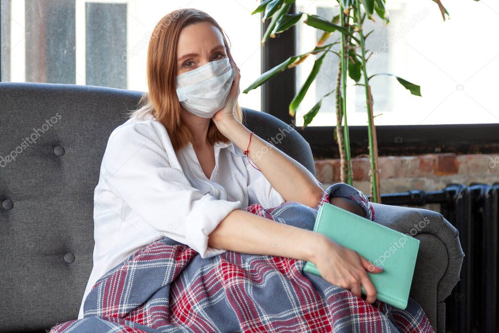 Woman in medical mask holding a book while sitting on a sofa