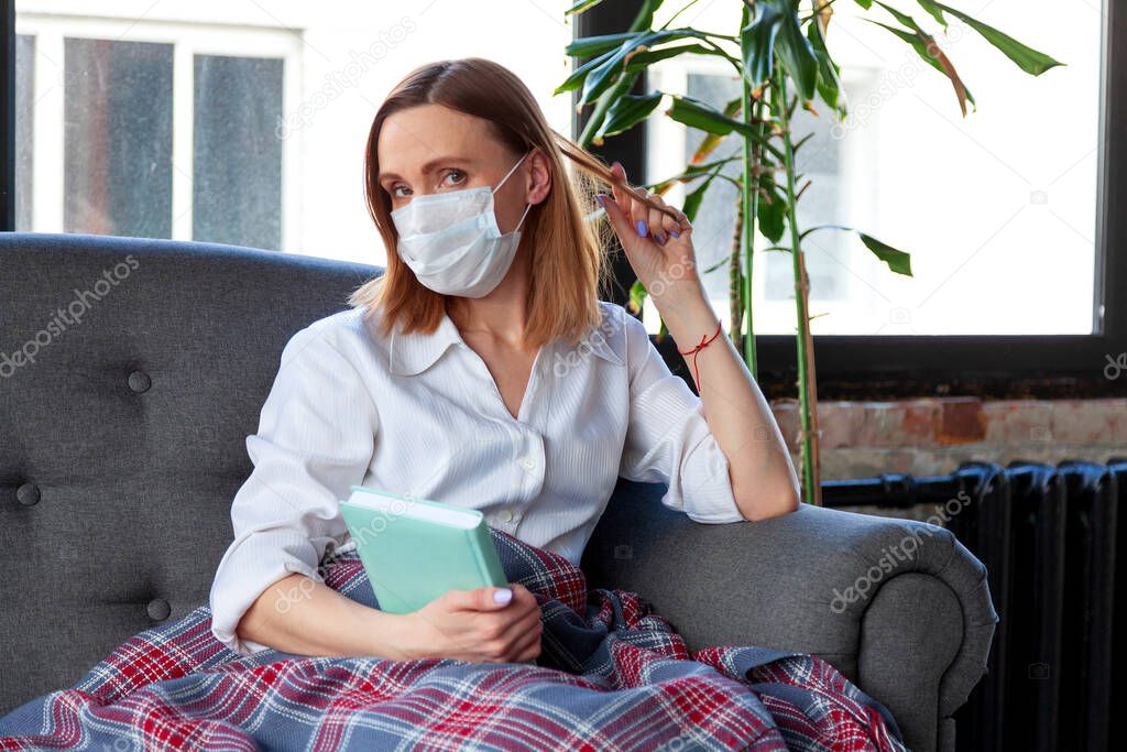 Woman in medical mask holding a book while sitting on a sofa