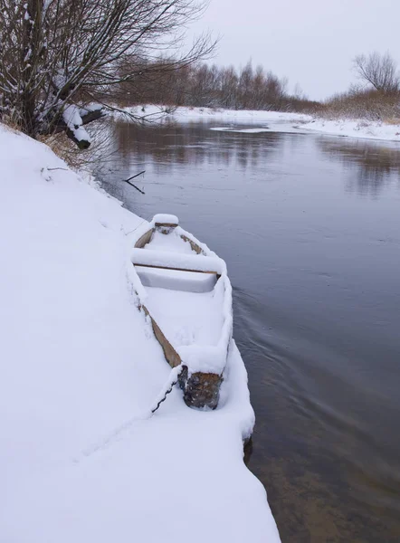 Bateau en bois recouvert de neige sur le rivage de la rivière d'hiver Photos De Stock Libres De Droits