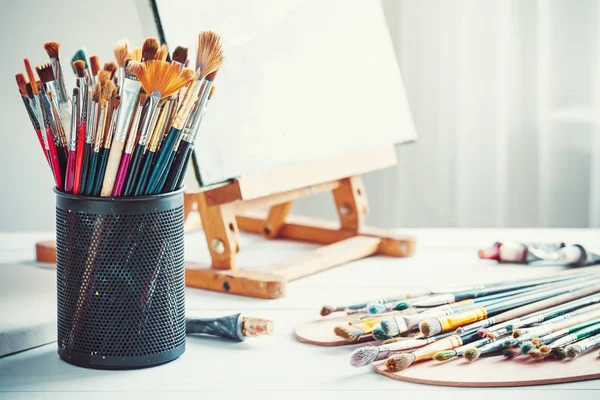 Artistic equipment: easel, paint brushes, tubes of paint, palette and paintings on work table in a artist studio. — Stock Photo, Image