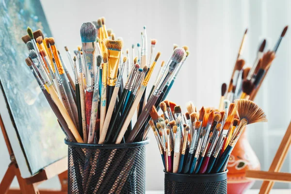 Artistic equipment in studio: wooden easel,  paintbrushes, tubes of paint, palette and paintings. — Stock Photo, Image