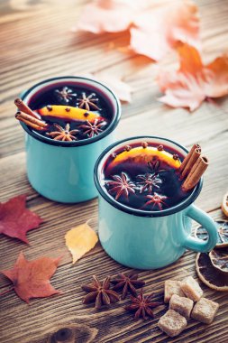 Mulled wine in blue enameled rustic mugs with spices and citrus  fruit on wooden table with autumn leaves. Retro toned clipart