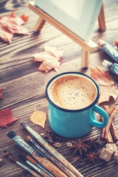Coffee cup, artist paintbrushes, paints, artistic canvas on easel and autumn leaves on desk — Stock Photo, Image
