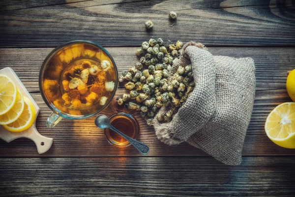 Healthy chamomile tea cup, hessian bag of dry daisy flowers, honey jar and lemon slices. Top view. — Stock Photo, Image