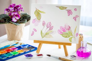 Canvas on easel, watercolor sketch, paints, brushes and violet senpolia flower in pot. clipart
