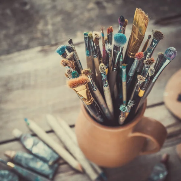 Paintbrushes in a jug from potters clay, wooden palette and paints. — Stock Photo, Image
