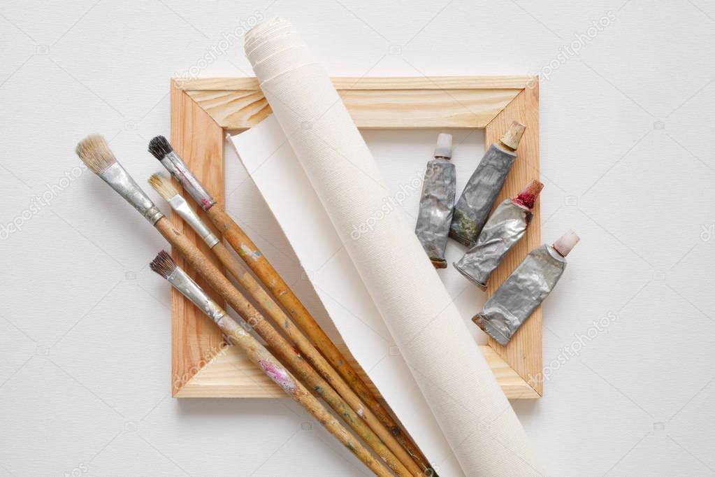 Wooden stretcher bar, paintbrushes, roll of artist canvas and paint tubes on white canvas background. Top view.