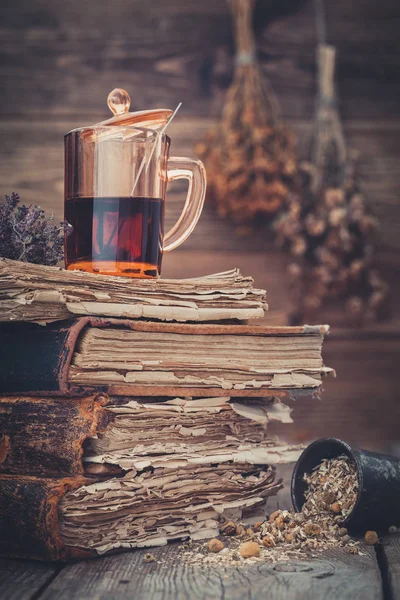 Xícara de chá saudável ou tintura de ervas na pilha de livros antigos, argamassa de ervas margaridas. Pendurado cachos de ervas medicinais no fundo . — Fotografia de Stock