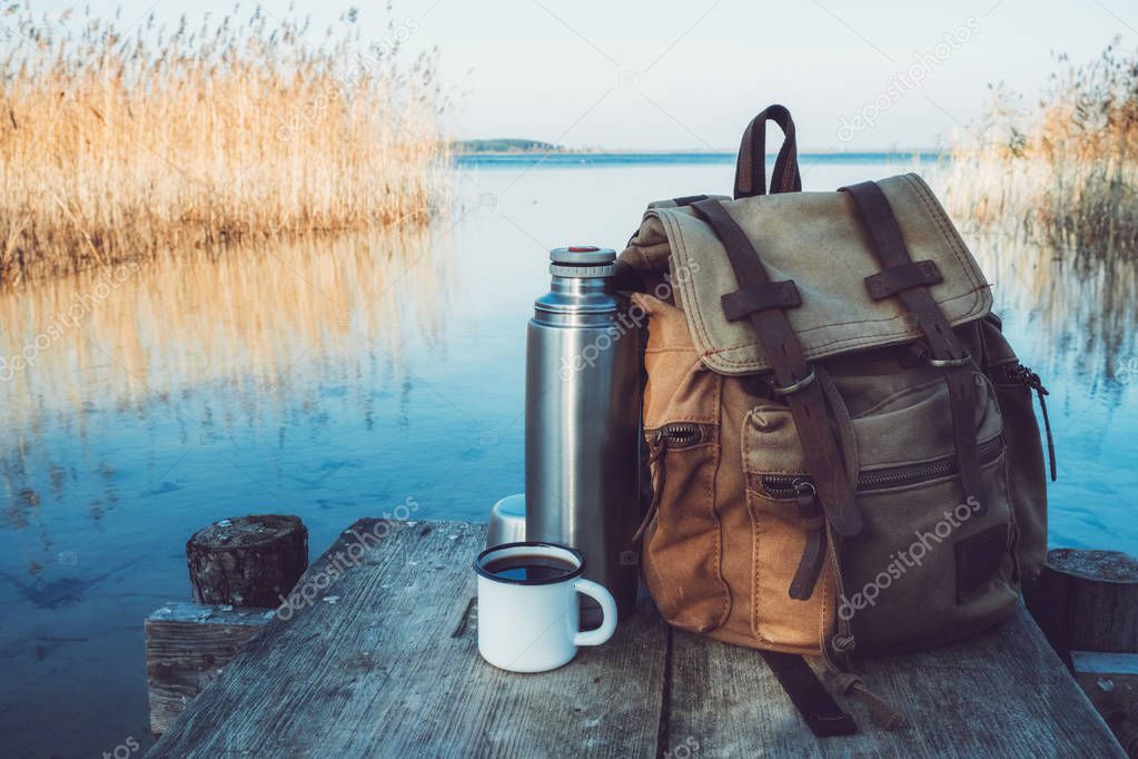 Enameled mug of coffee or tea, backpack of traveller and thermos