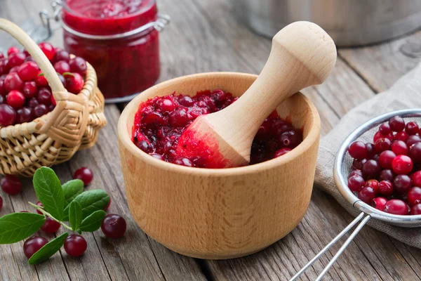 Houten kom van geplette veenbessen, mand van moerbeien, pot van geplette bessen, jam of saus en zeef van veenbessen op de voorgrond. — Stockfoto