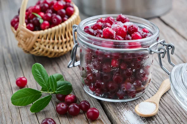 Jar of ripe cranberries with sugar, basket of bog berries and saucepan on background. — Stock Photo, Image