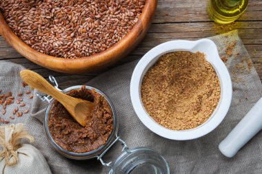 Glass jar of raw organic paste from flax seed, mortar of linseeds crushed into powder, wooden bowl of whole flax seeds and oil bottle on table. clipart