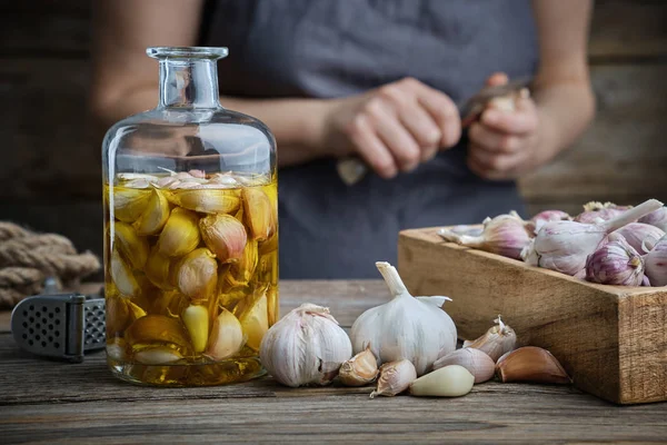 Garrafa de óleo ou infusão aromática de alho e caixa de madeira de dentes de alho na mesa de cozinha de madeira. Mulher descasca alho com uma faca no fundo . — Fotografia de Stock