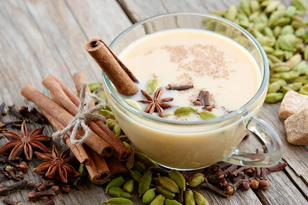 Cup of healthy masala tea with milk and aromatic spices and herbs. Ayurveda treatments. Cinnamon sticks, cardamom, allspices and anise on wooden table. — Stock Photo, Image