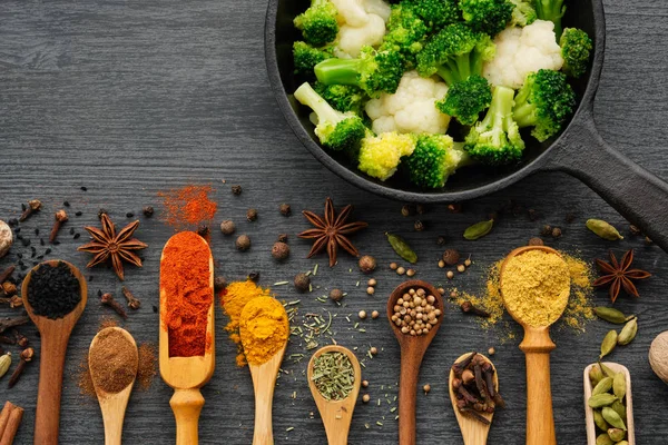 Diverse aromatische kleurrijke specerijen en kruiden in houten lepels en schepjes. Broccoli en bloemkool op ijzeren koekenpan. — Stockfoto