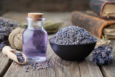 Kuru lavanta çiçekleri kasede ve bir şişe lavanta yağı ya da eritilmiş su. Arka planda eski kitaplar ve lavanta çiçekleri var.. 