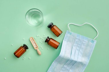 Bir şişe homeopatik granül, tıbbi çözelti hazırlamak için su bardağı, yeşil arka planda koruyucu tıbbi maske. Yukarıdan görüntüle.