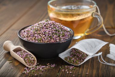 Bowl of dry healthy heather, tea bag with Erica flowers. Glass tea cup with herbal teabag inside on wooden table. Herbal medicine.  clipart
