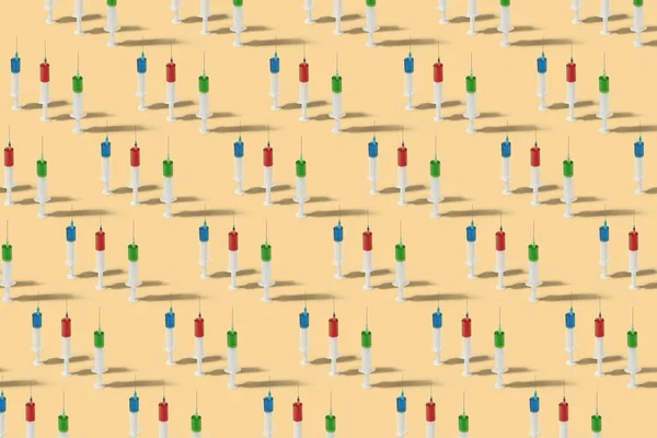 Medicinal pattern from disposable vertical standing plastic syringes with red, green and blue vaccine or serum for injection on a sand color background, with shadows.