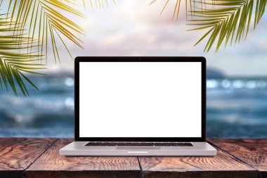 Ahşap bir masada, yeşil palmiye yapraklı bulanık deniz manzaralı boş bir laptop monitörü, kopyalama alanı. Uzaktan çalışma kavramı.