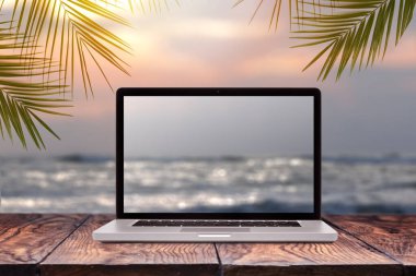Notebook monitor with blurred seascape sunrise picture on a wooden table against the same background with green palm leaves frame, copy space. Working at sea, outside office concept. clipart