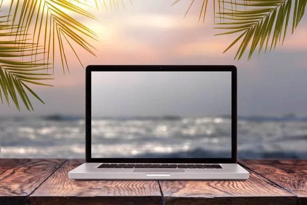 Notebook monitor with blurred seascape sunrise picture on a wooden table against the same background with green palm leaves frame, copy space. Working at sea, outside office concept.