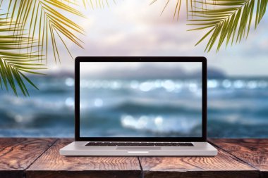 Laptop monitor with blurred seascape image on a wooden table against the same background with green palm leaves frame, copy space. Working remotely at seacoast, on nature outside office concept. clipart