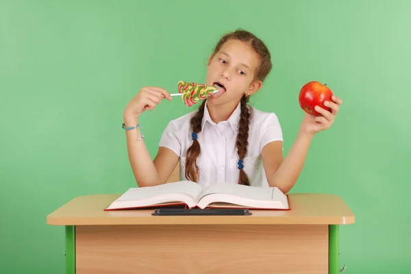 Девушка в школьной форме сидит за столом и выбирает конфеты или яблоко — стоковое фото