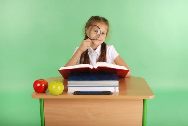 Girl in a school uniform sitting at a desk with a magnifying glass — Stock Photo, Image