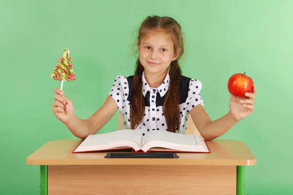 Lány az iskolában egységes ül az asztalon, és válassza ki a candy, vagy egy Alma — Stock Fotó