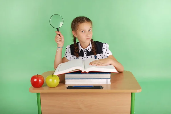 Menina em um uniforme escolar sentado em uma mesa com uma lupa — Fotografia de Stock