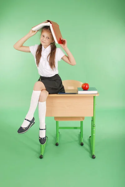 Education, people, children and school concept - young school girl sitting on desk with a book on her head — Stock Photo, Image