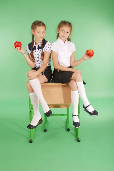 Two girls in a school uniform sitting on desk and eat apples — Stock Photo, Image