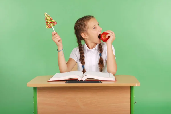 Девушка в школьной форме сидит за столом и выбирает конфеты или яблоко — стоковое фото
