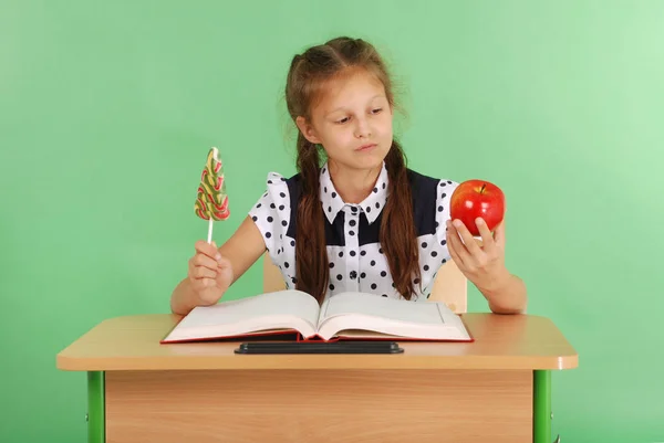 Lány az iskolában egységes ül az asztalon, és válassza ki a candy, vagy egy Alma — Stock Fotó