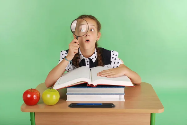 Girl in a school uniform sitting at a desk with a magnifying glass — Stock Photo, Image