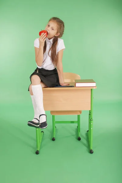 Девушка в школьной форме сидит на столе и ест яблоко — стоковое фото