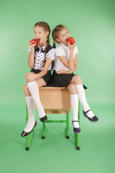 Two girls in a school uniform sitting on desk and eat apples — Stock Photo, Image