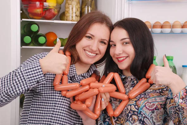 Two young girls holds sausages on the refrigerator background