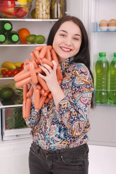 Молода дівчина тримає сосиски на фоні холодильника — стокове фото