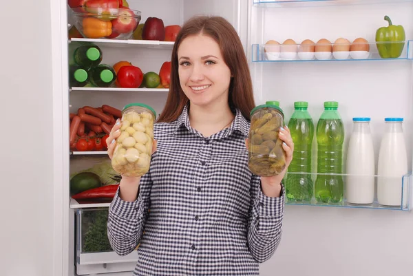 Chica joven sostiene champiñones enlatados y pepinos en el fondo del refrigerador — Foto de Stock